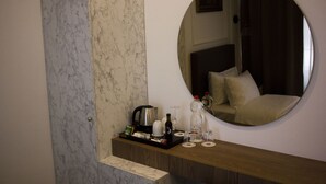 Ett hotellrum med en vägg av marmor, en rund spegel, ett litet träbord med en kaffebryggare, vinglas och en flaska vatten.
