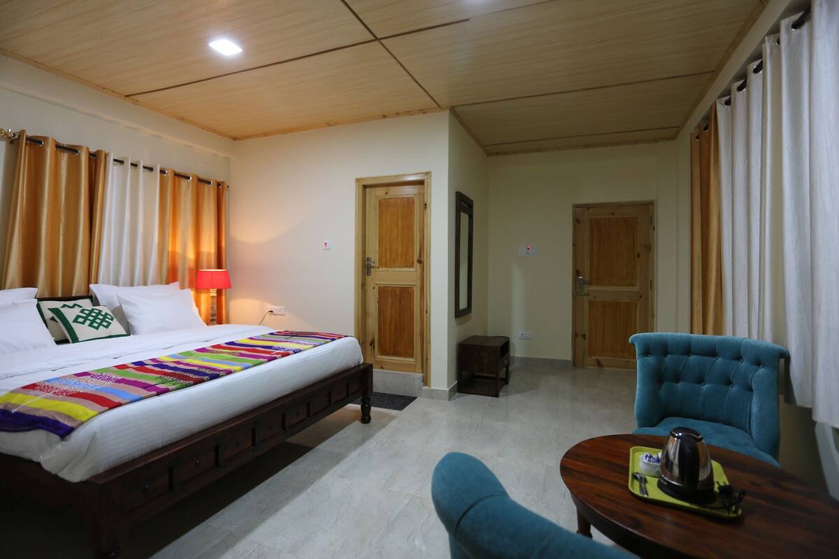 A hotel room with a bed, a chair, a small table with a teapot, and a door leading to another room.