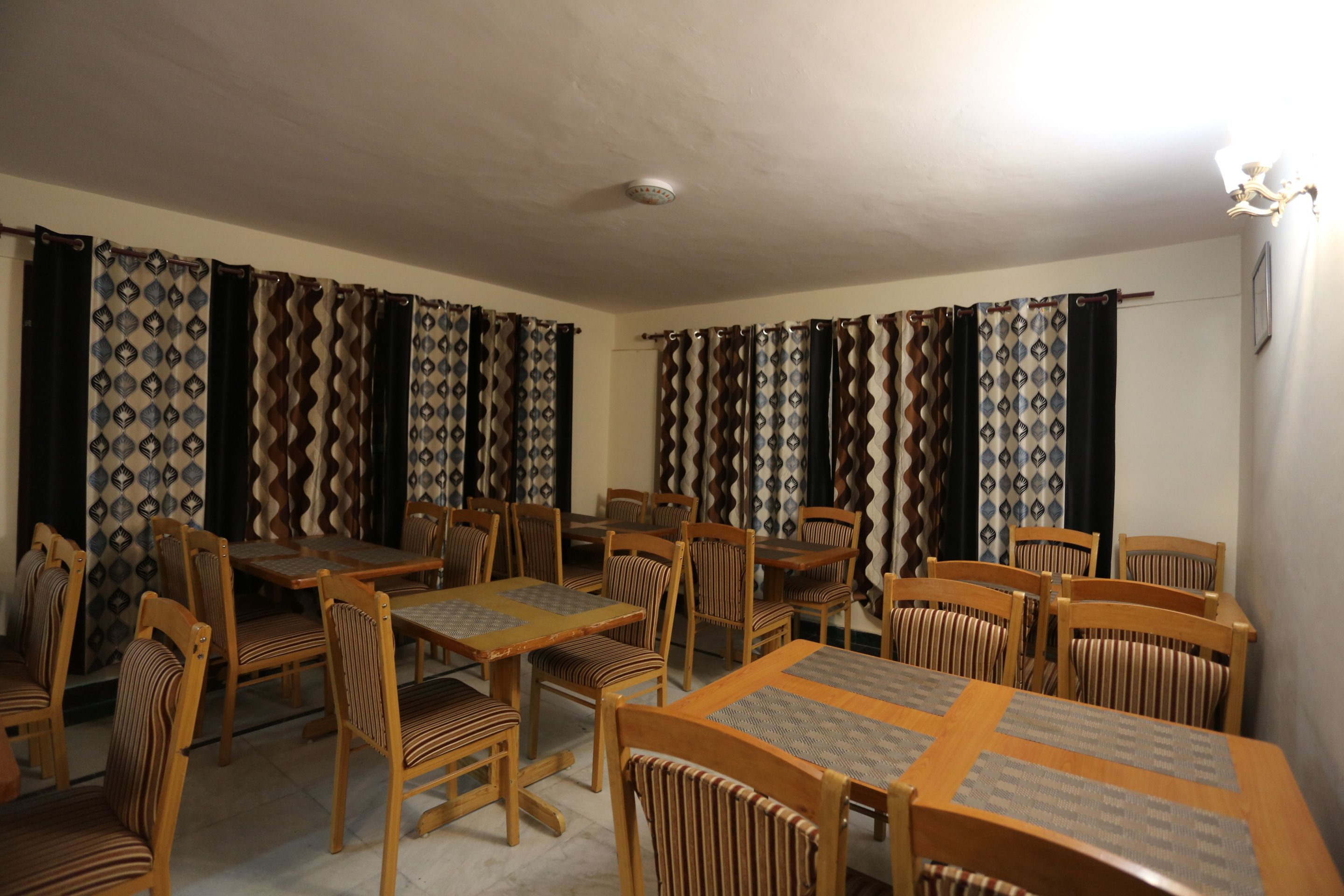 Un espace repas avec plusieurs tables et chaises en bois, des rideaux à motifs et un luminaire suspendu au plafond.