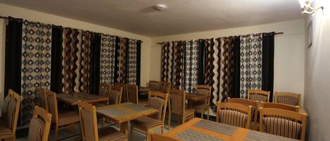 Un espace repas avec plusieurs tables et chaises en bois, des rideaux à motifs et un luminaire suspendu au plafond.