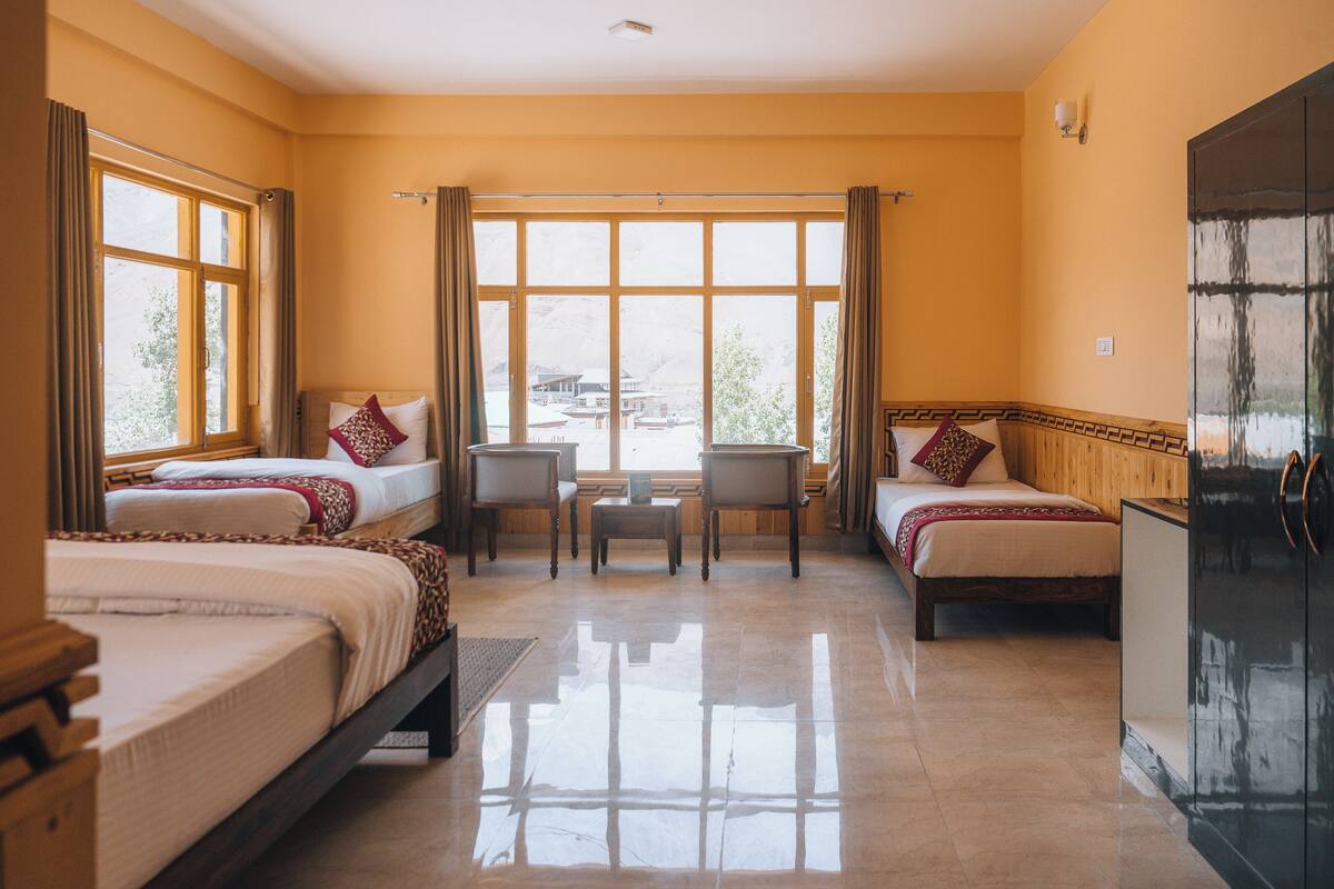 A hotel room with two beds, a desk, chairs, a refrigerator, and large windows with curtains.