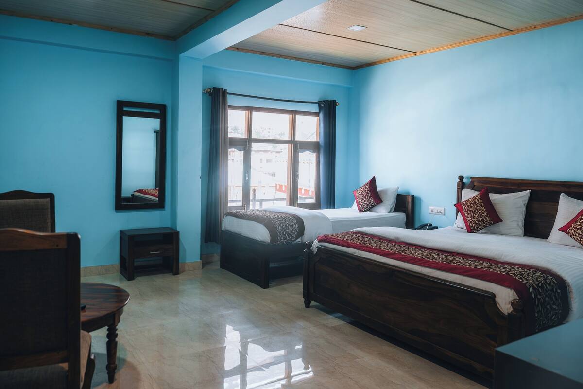 A bedroom with two beds, a wooden headboard, a small table, a chair, a mirror, and a window with curtains.
