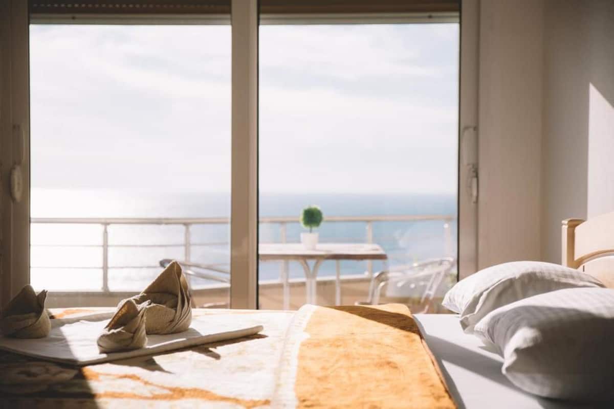 Une chambre d’hôtel avec un lit, une table agrémentée d’une plante et une vue sur la mer grâce à de grandes fenêtres.