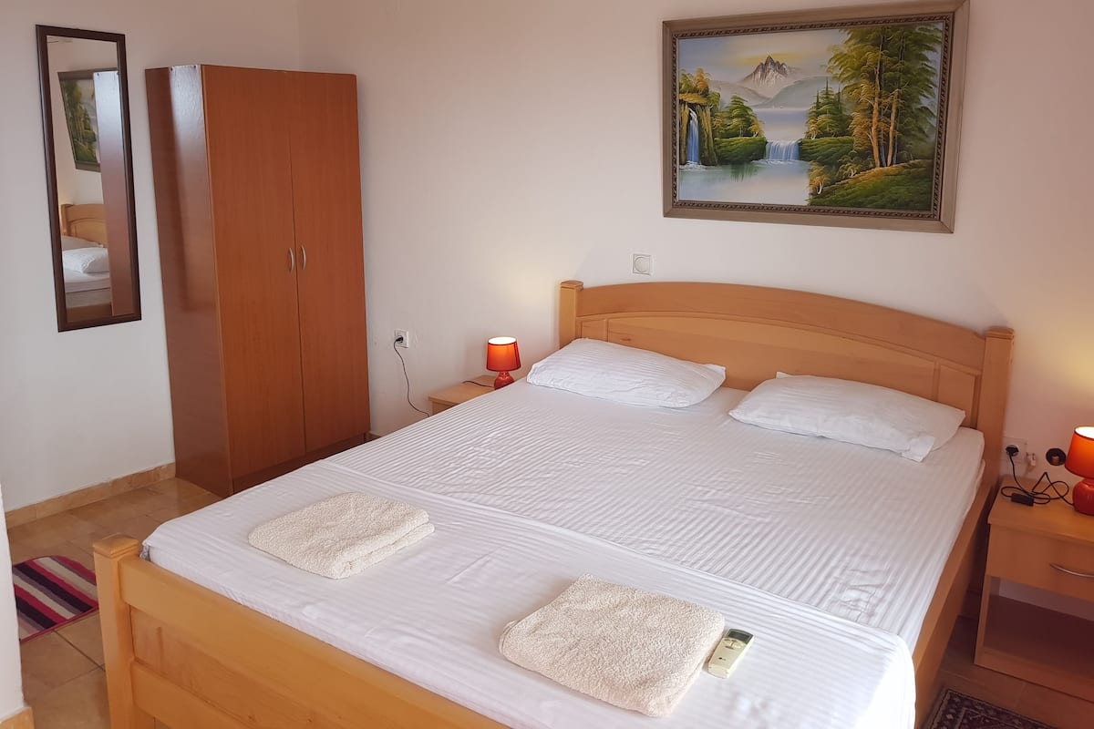 Un lit bien fait, avec du linge de lit blanc et deux serviettes, une tête de lit en bois, une table de chevet avec une lampe, une armoire et un tableau représentant un paysage accroché au mur.