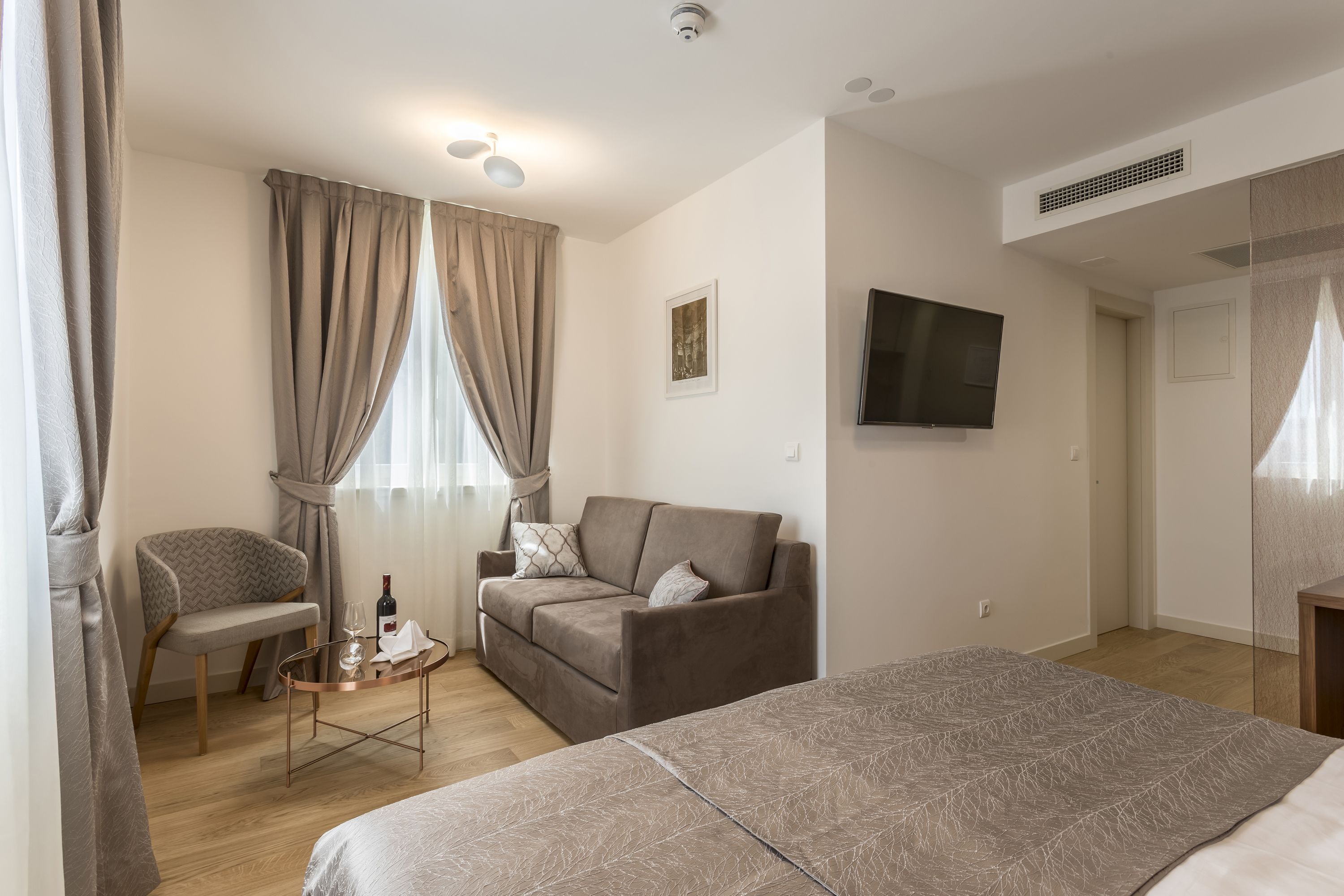 A modern hotel room with a bed, a sofa, a chair, and a small table with a bottle and glass.