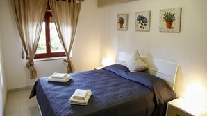 Un dormitorio con una cama, una ventana con cortinas, una mesita de noche con lámpara y tres cuadros colgados en la pared con motivos vegetales.