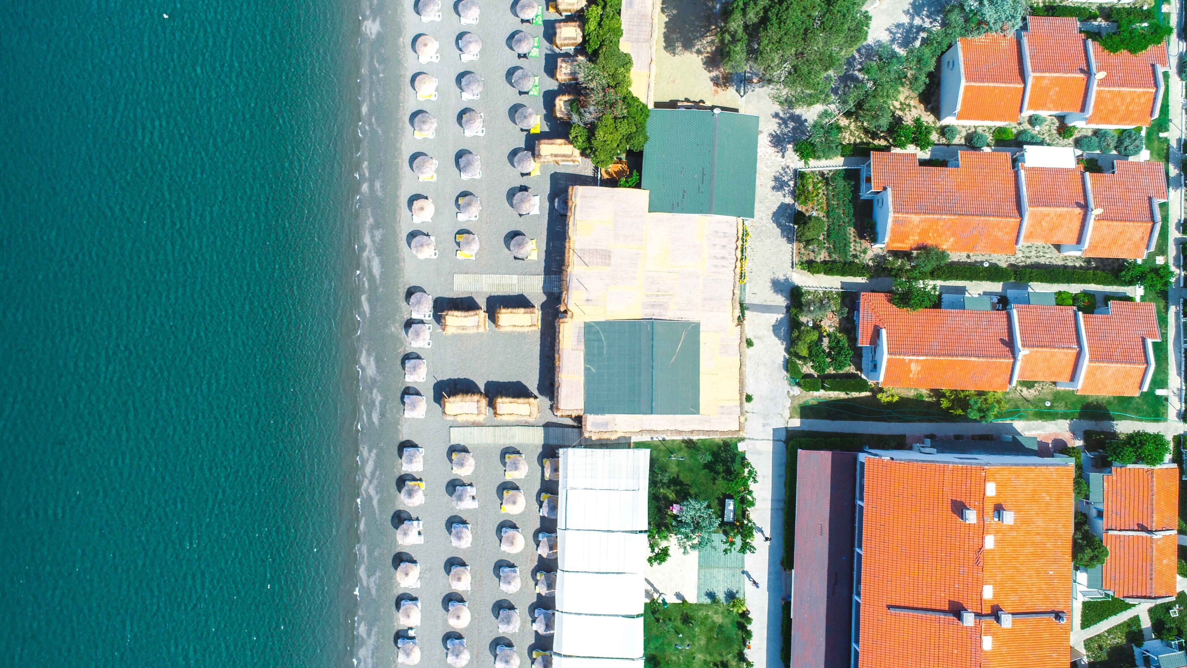 An aerial view of a coastal area with a beach, rows of beach umbrellas, and adjacent buildings with red-tiled roofs.