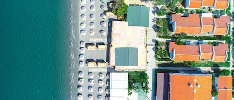An aerial view of a coastal area with a beach, rows of beach umbrellas, and adjacent buildings with red-tiled roofs.