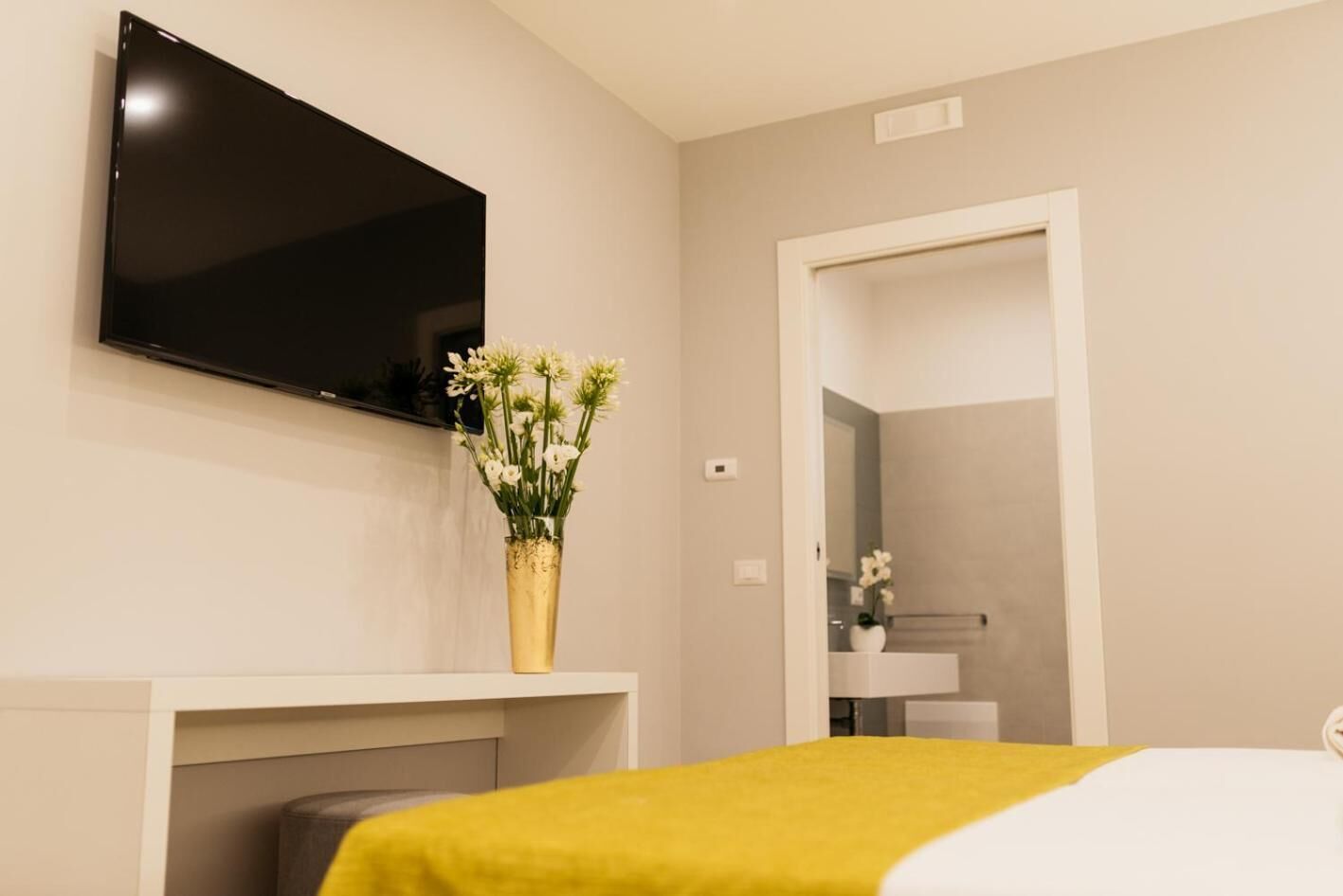 Habitación de hotel con cama, televisor montado en la pared, un jarrón con flores y un baño visible a través de una puerta abierta.