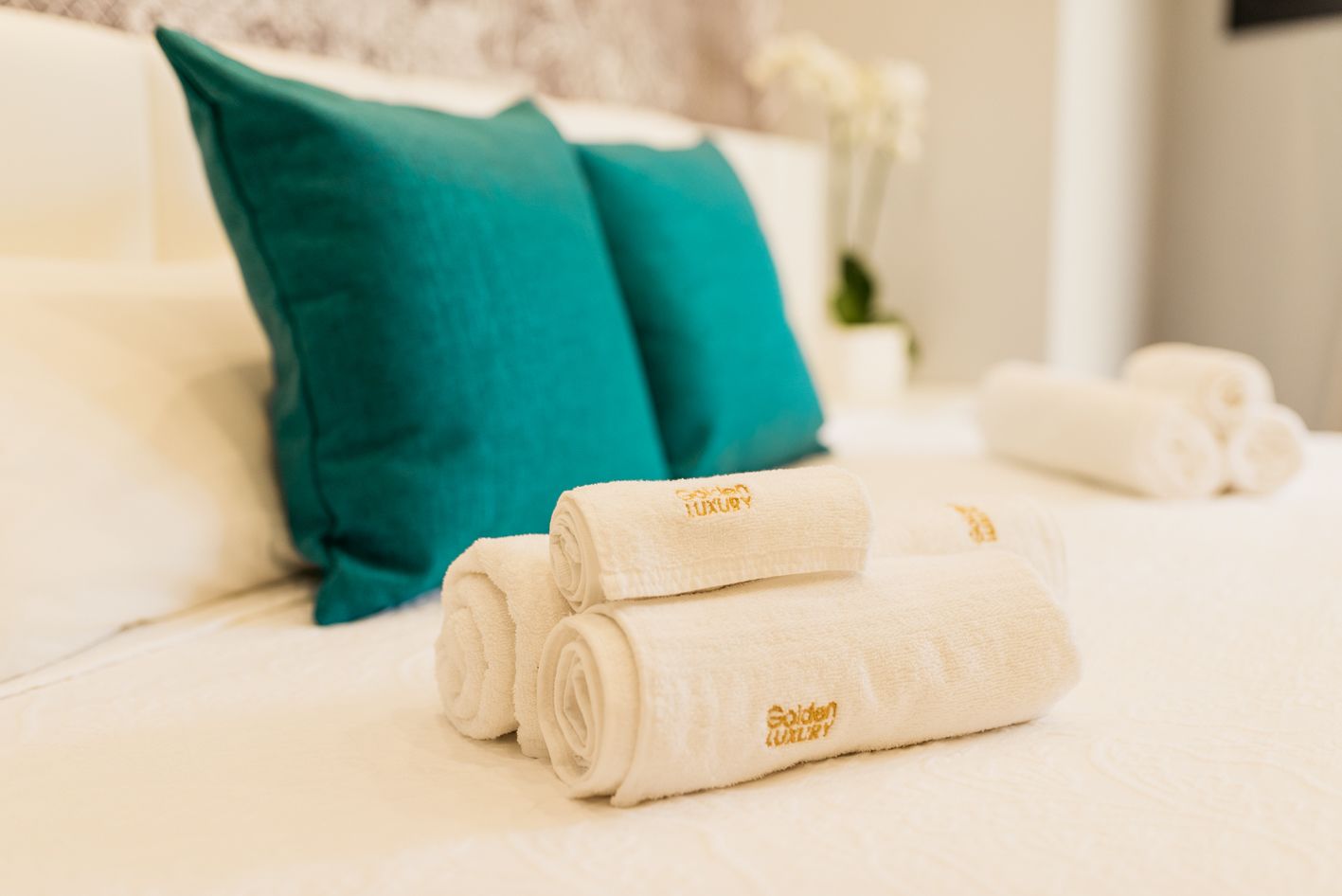 Una cama bien hecha con almohadas blancas y turquesas, y toallas blancas enrolladas con un logotipo.