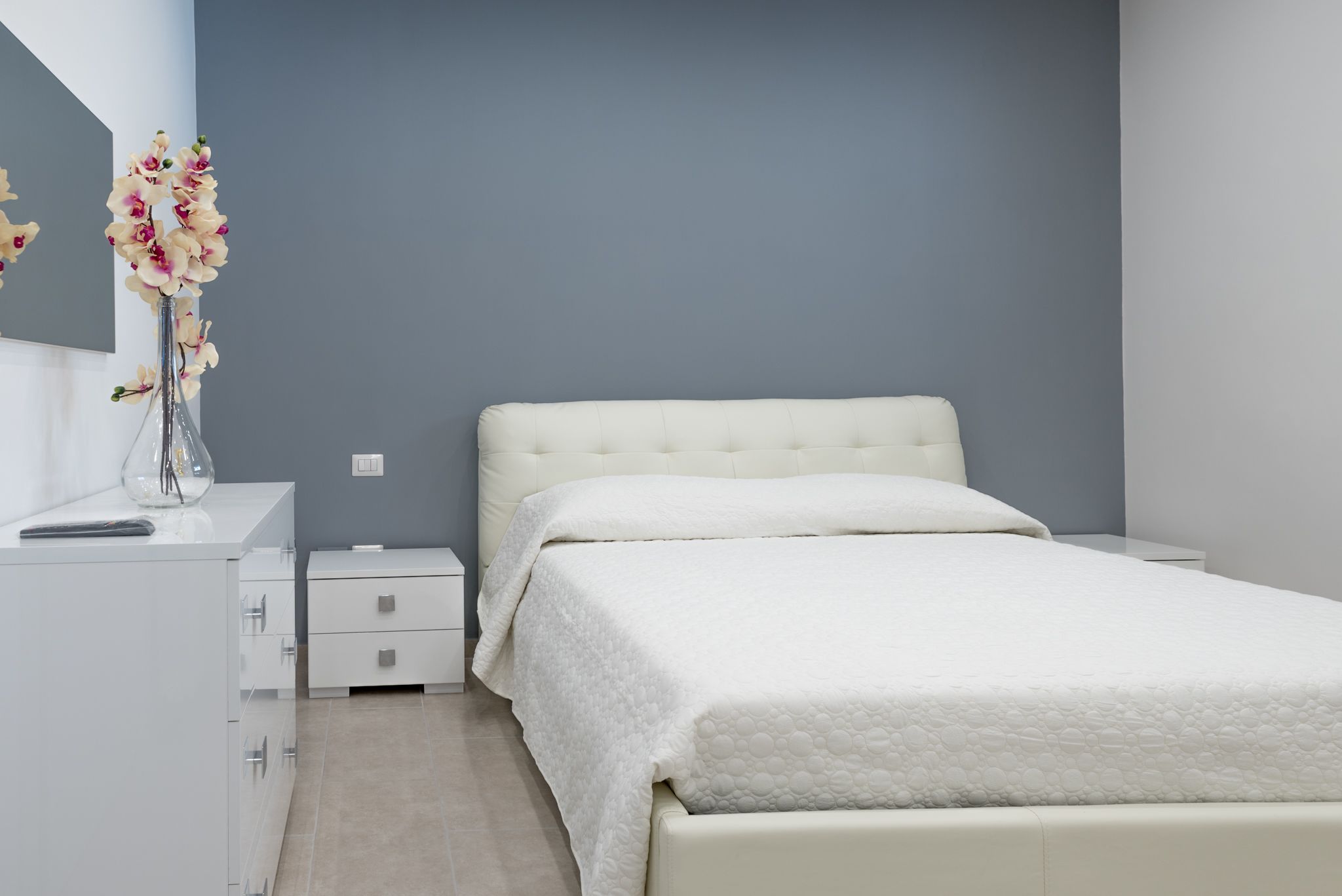 Un dormitorio moderno con una cama grande, una mesita de noche, un aparador y un florero con flores.