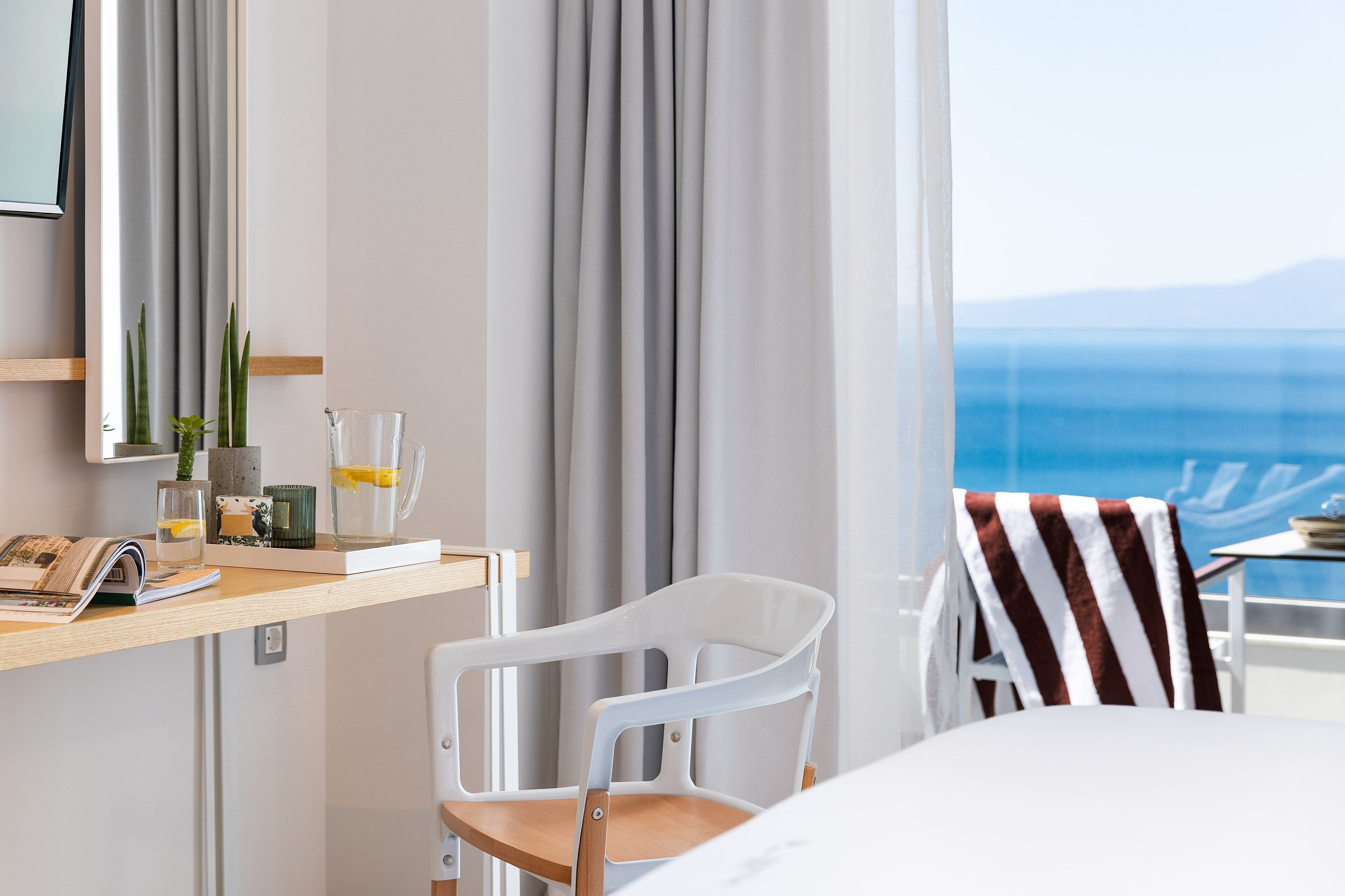 Een hotelkamer met een houten bureau, een witte stoel, een gestreepte handdoek en uitzicht op zee.