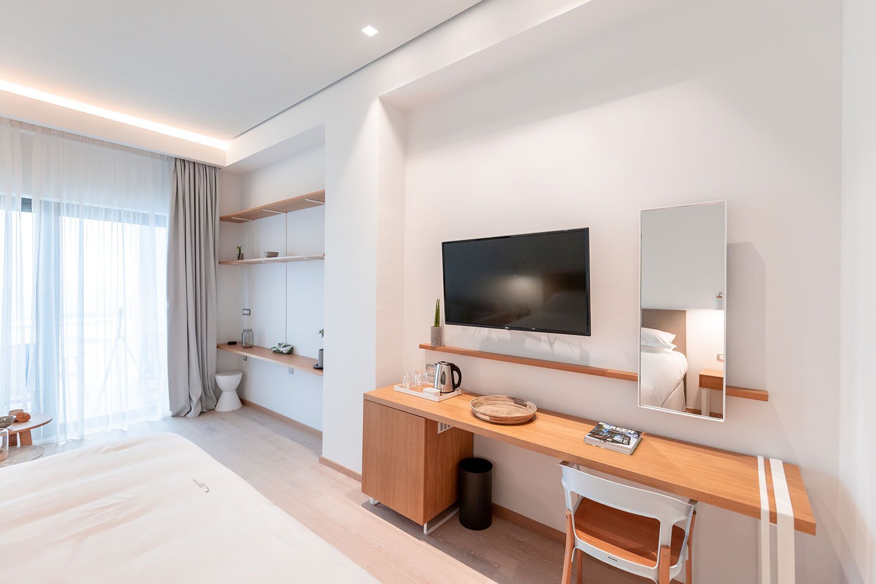 A modern hotel room with a large bed, a desk with a mirror, a television, and built-in shelves.