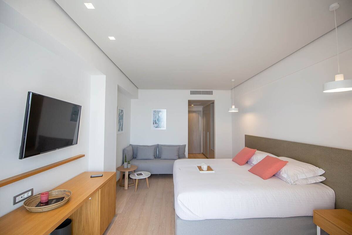 A modern hotel room with a flat-screen TV, a bed with white bedding and red pillows, a grey sofa, a small round table with a cup and a book, and a wooden desk with a basket and a remote control.
