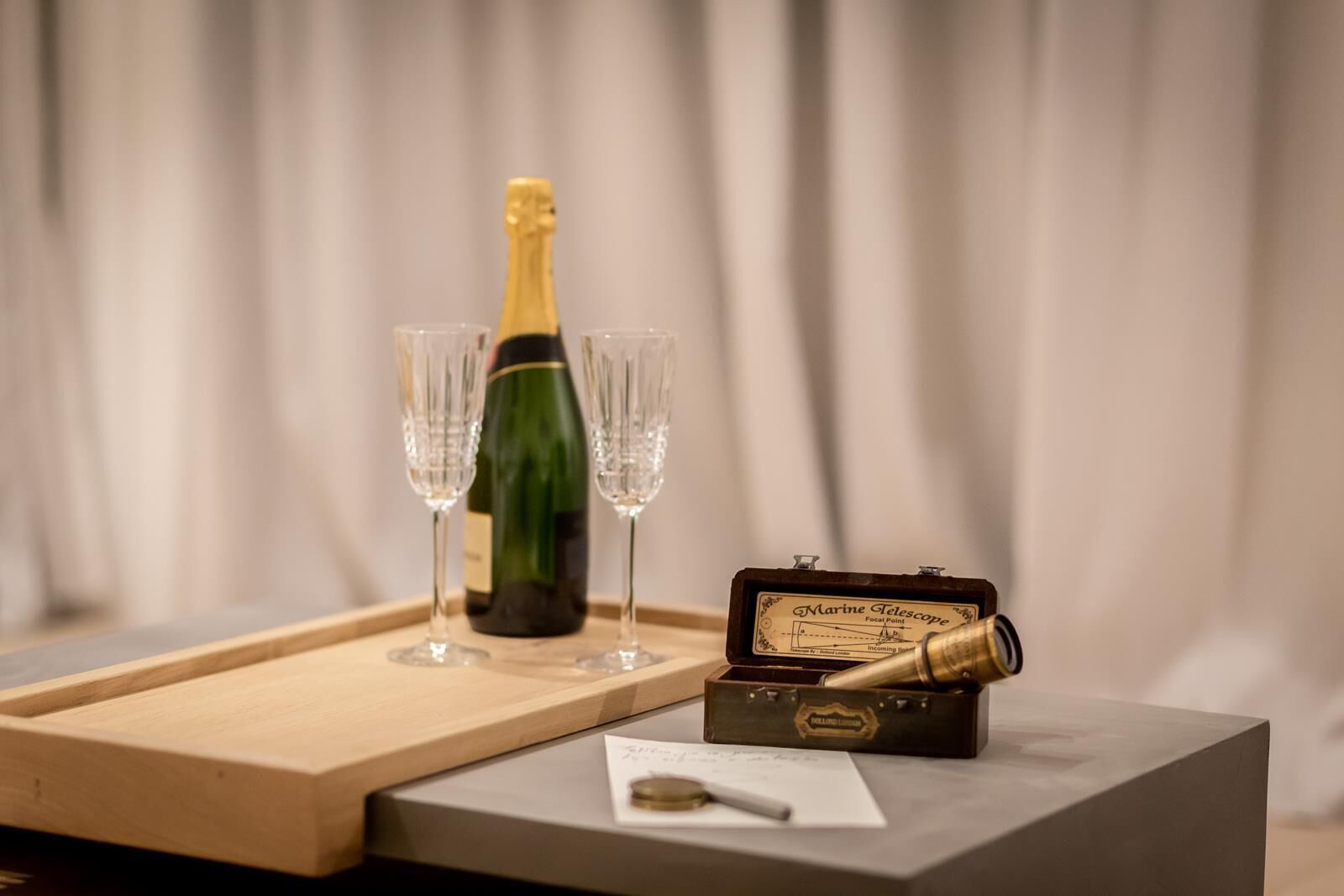 A wooden tray with a bottle of champagne and two glasses, a marine telescope, and a small box with a label.