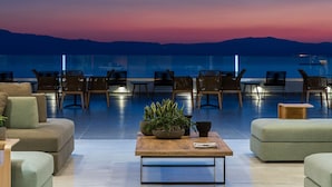 Un espace de détente moderne, doté de canapés, de fauteuils et d’une table basse, offrant une vue sur la mer et les montagnes au crépuscule.