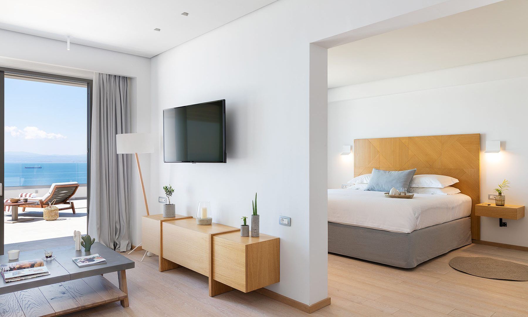 A modern hotel room with a large bed, a TV mounted on the wall, a balcony with a view of the sea, and a small desk with a plant.