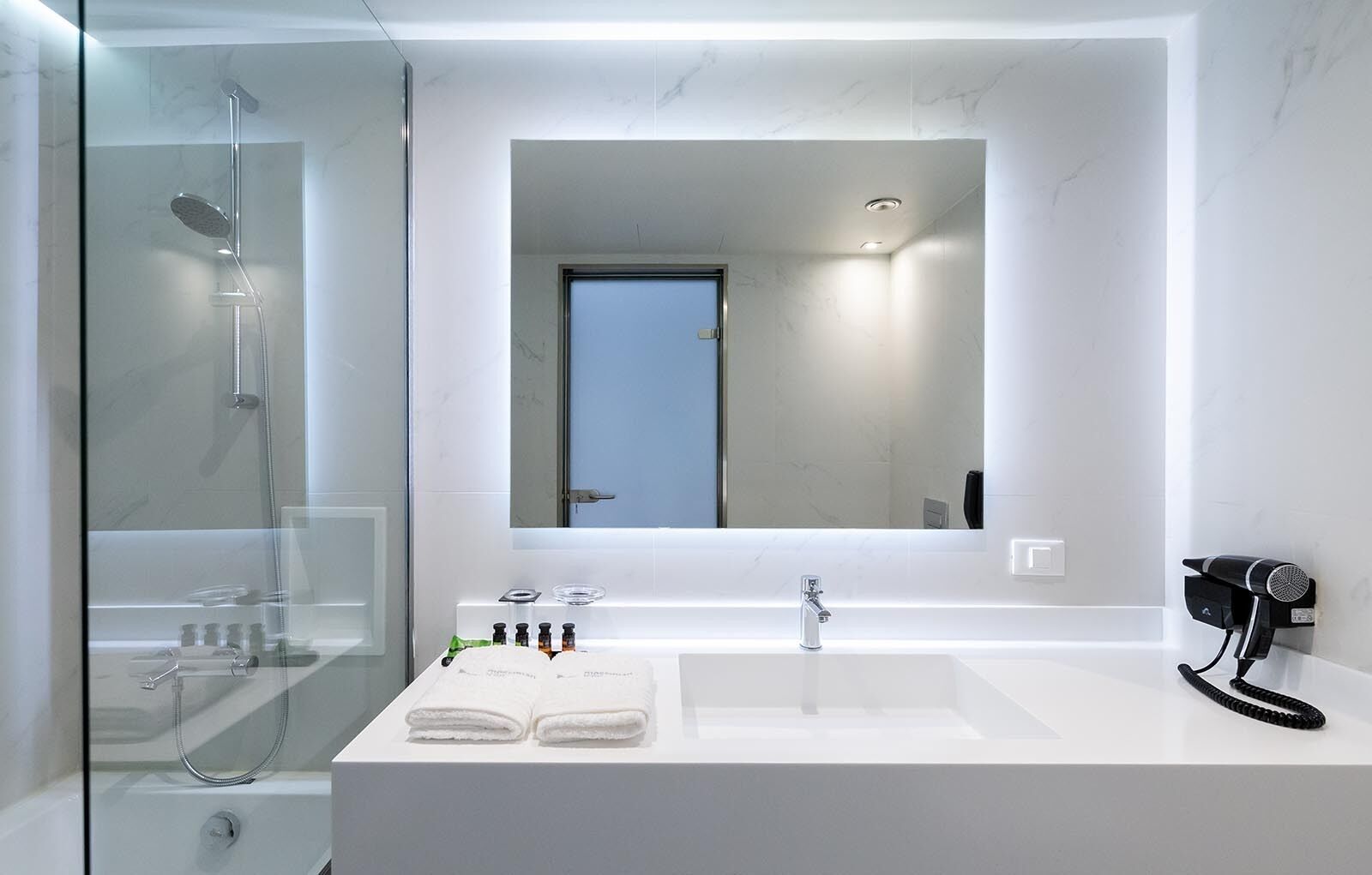 A modern bathroom with a glass-enclosed shower, a white vanity with a large mirror, and a hairdryer mounted on the wall.