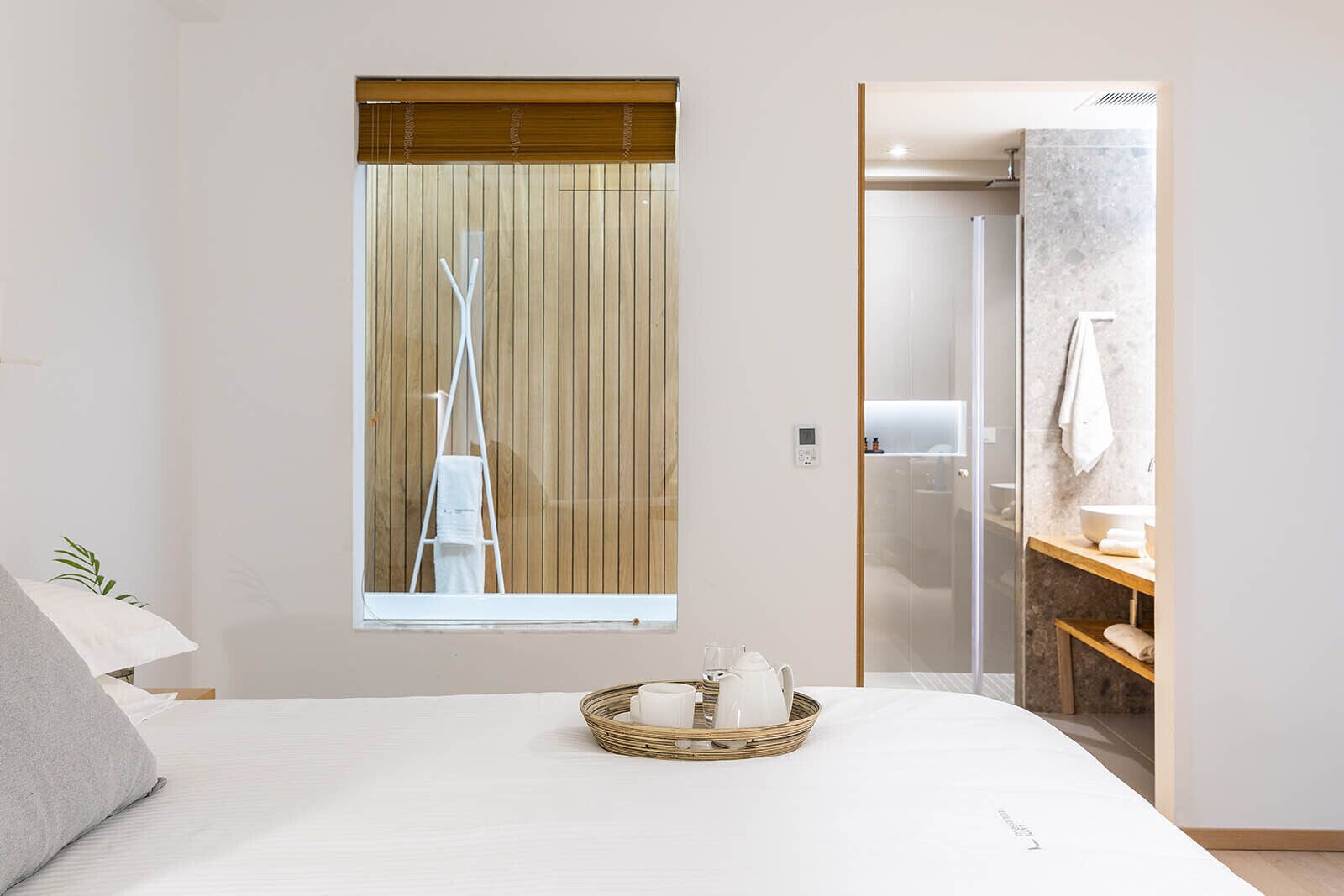 A modern bedroom with a large bed, a window with blinds, a bedside table with a tray of toiletries, and a bathroom visible through an open door.