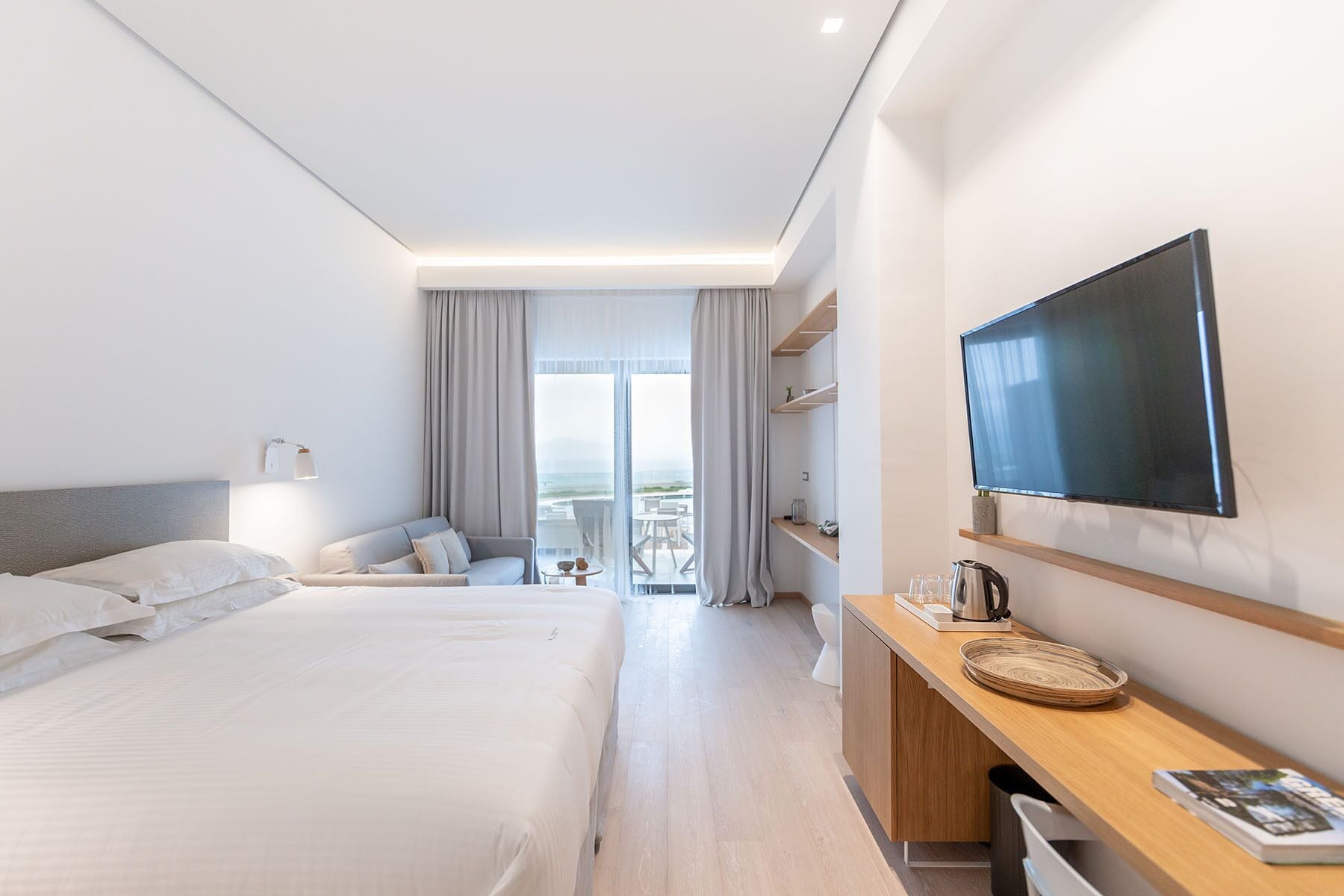 Ein modernes Hotelzimmer mit einem großen Bett, einem an der Wand montierten Fernseher, einem Schreibtisch mit Wasserkocher und einem Balkon mit Blick.