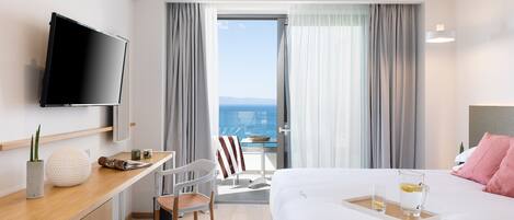 A modern hotel room with a bed, a desk with a flat-screen TV, a chair, a small table with a tray holding a glass of water and a pitcher, and a view of the sea through large windows.