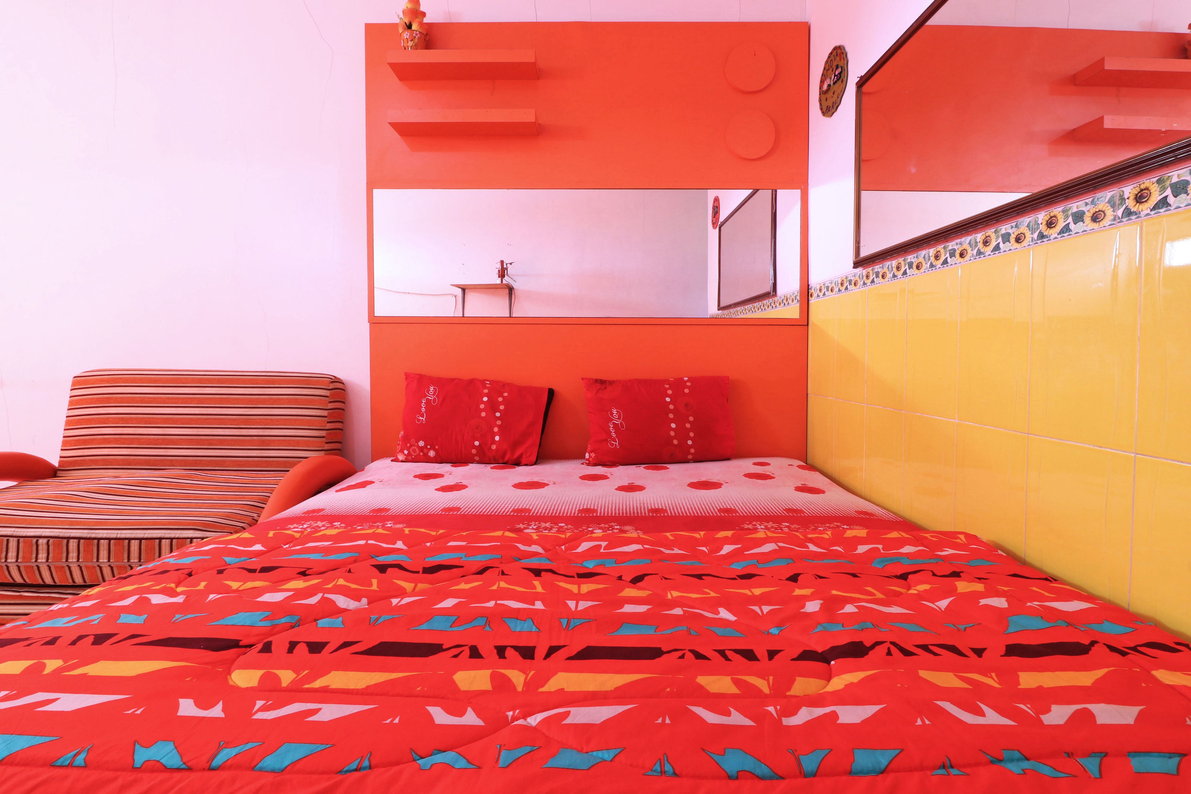 A bed with a colorful patterned bedspread, a striped armchair, and a bright orange headboard with a mirror.