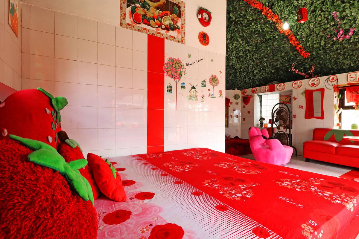 A room with a large bed, a pink chair, and a red sofa. There is a wall with fruit and vegetable decorations, and a green leafy ceiling.