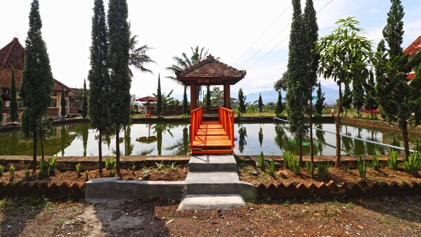 A wooden bridge leading to a gazebo over a pond, surrounded by trees and buildings. - Hotel Agung Permata Artha (Garut)