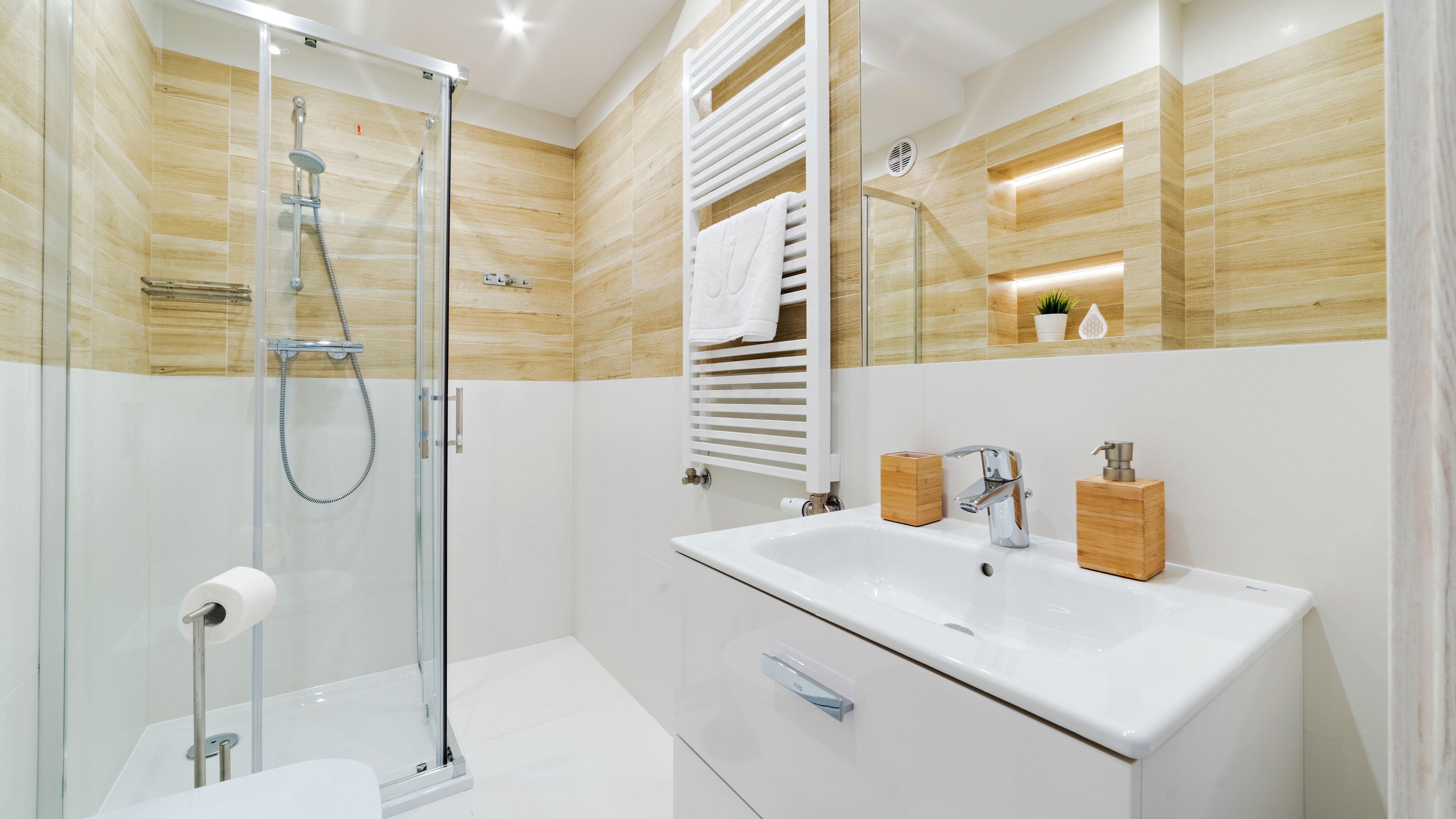 Un baño moderno con cabina de ducha de vidrio, un lavabo blanco y un dispensador de jabón de madera, y un perchero con una toalla blanca.