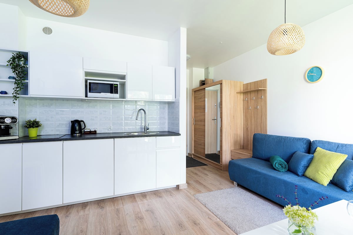 A modern kitchen with white cabinets, a microwave, and a coffee maker. There is a blue sofa with cushions and a red candle in the living area.