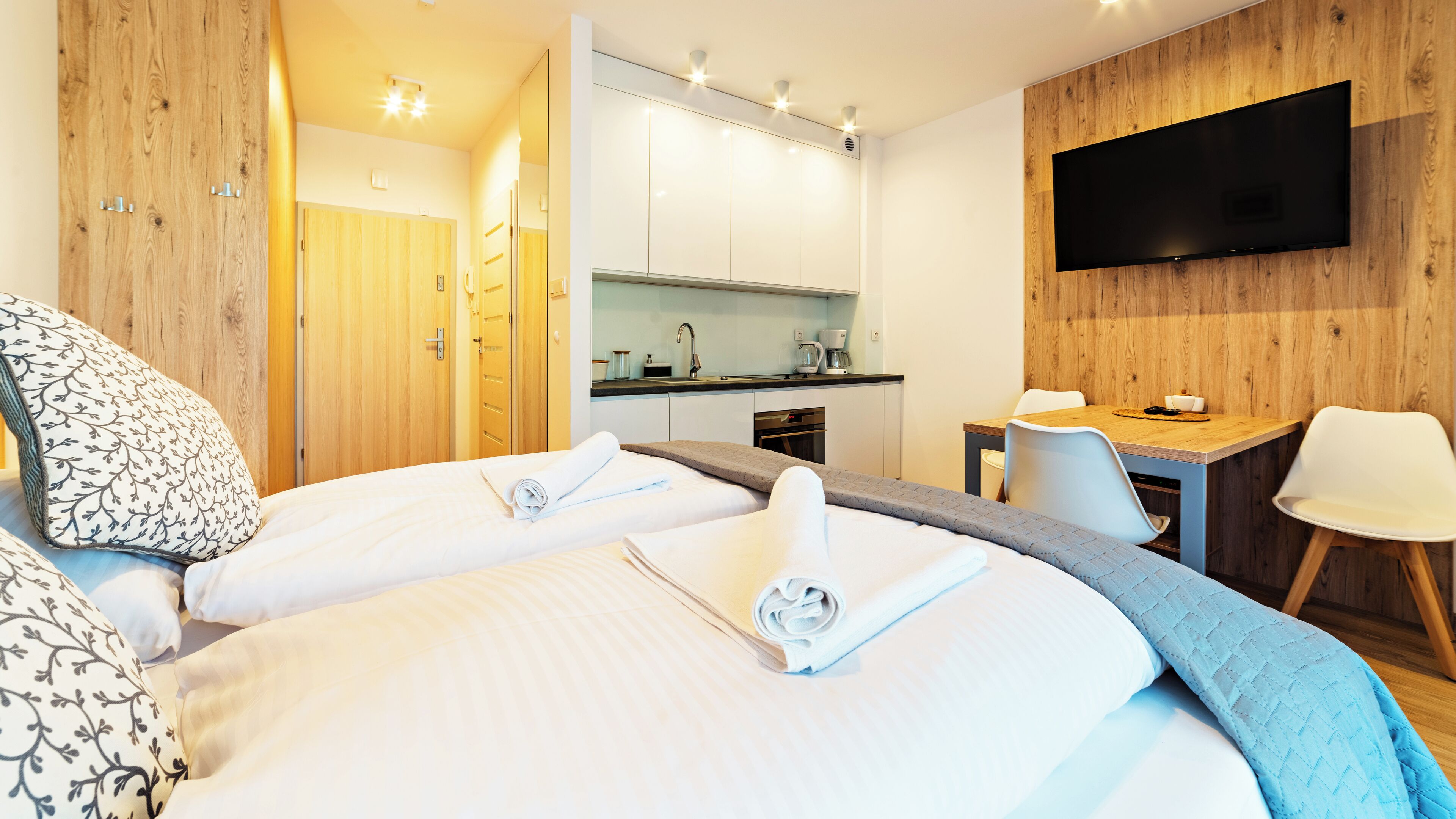 Habitación de hotel con dos camas, kitchenette, televisor y escritorio con sillas.