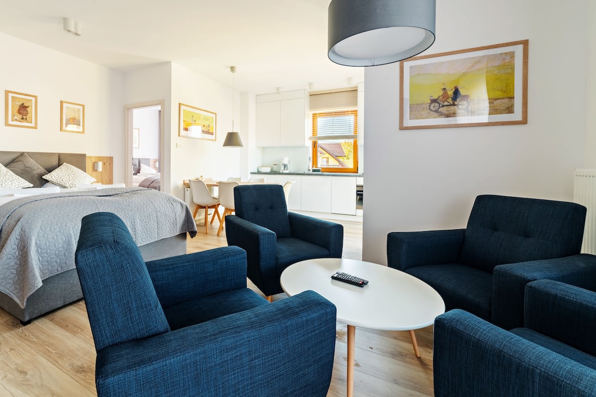 A modern hotel room with a bed, blue armchairs, a white table, and a kitchen area in the background.