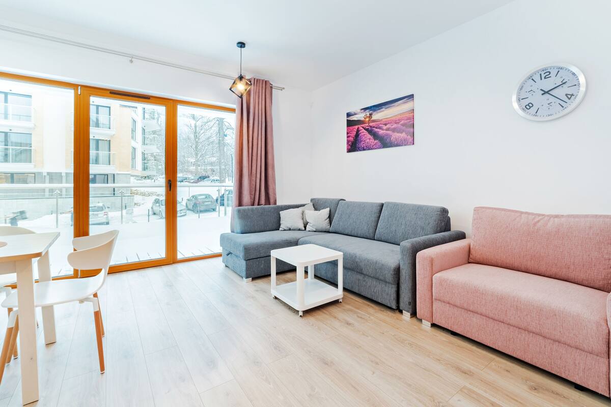 A modern living room with a grey sofa, a pink armchair, and a white coffee table. There is a dining area with a white table and chairs. A large window offers a view of the outside.