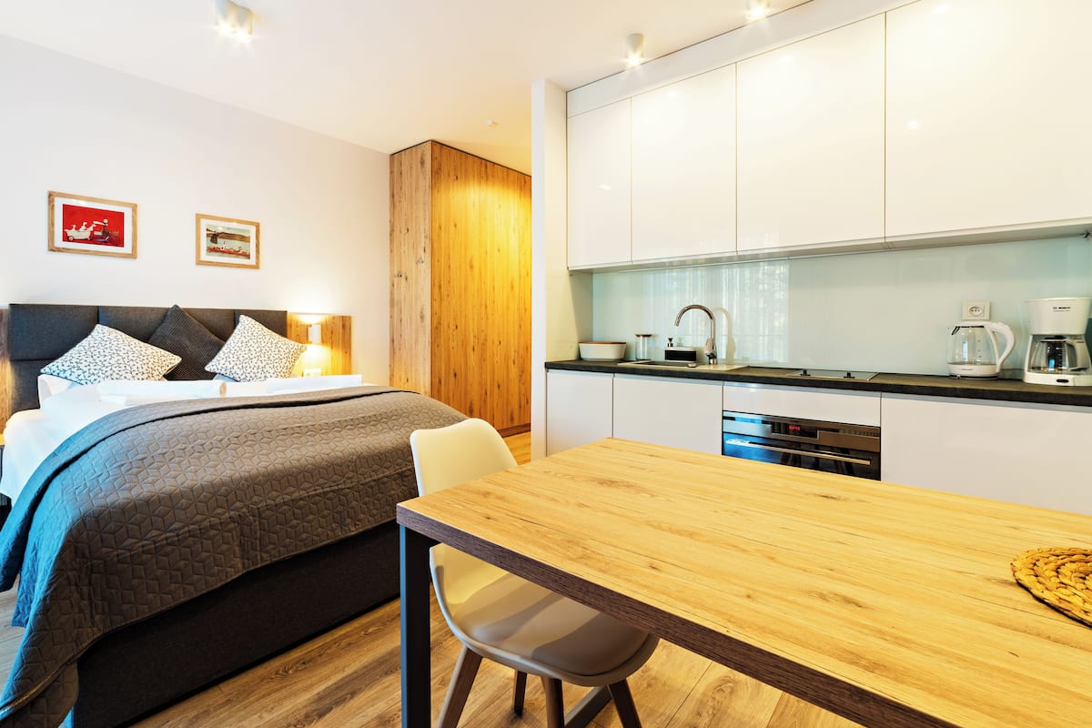 A modern hotel room with a bed, bedside tables, a desk, and a kitchenette.
