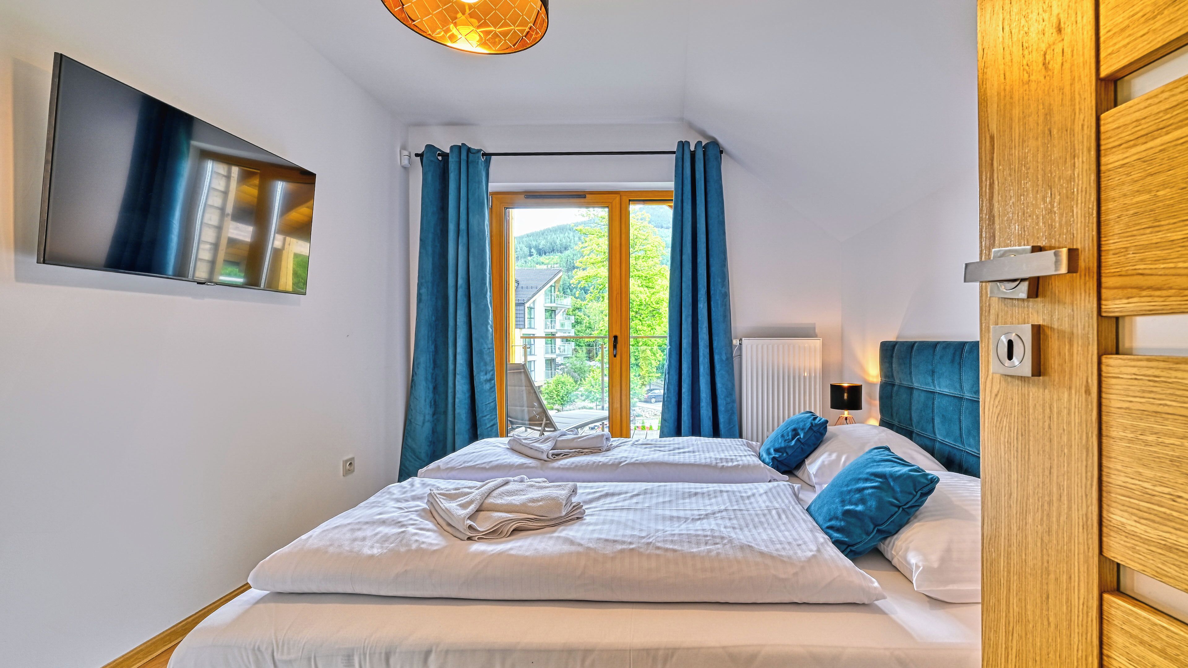 A bedroom with a bed, a TV mounted on the wall, a window with curtains, and a view of greenery outside.
