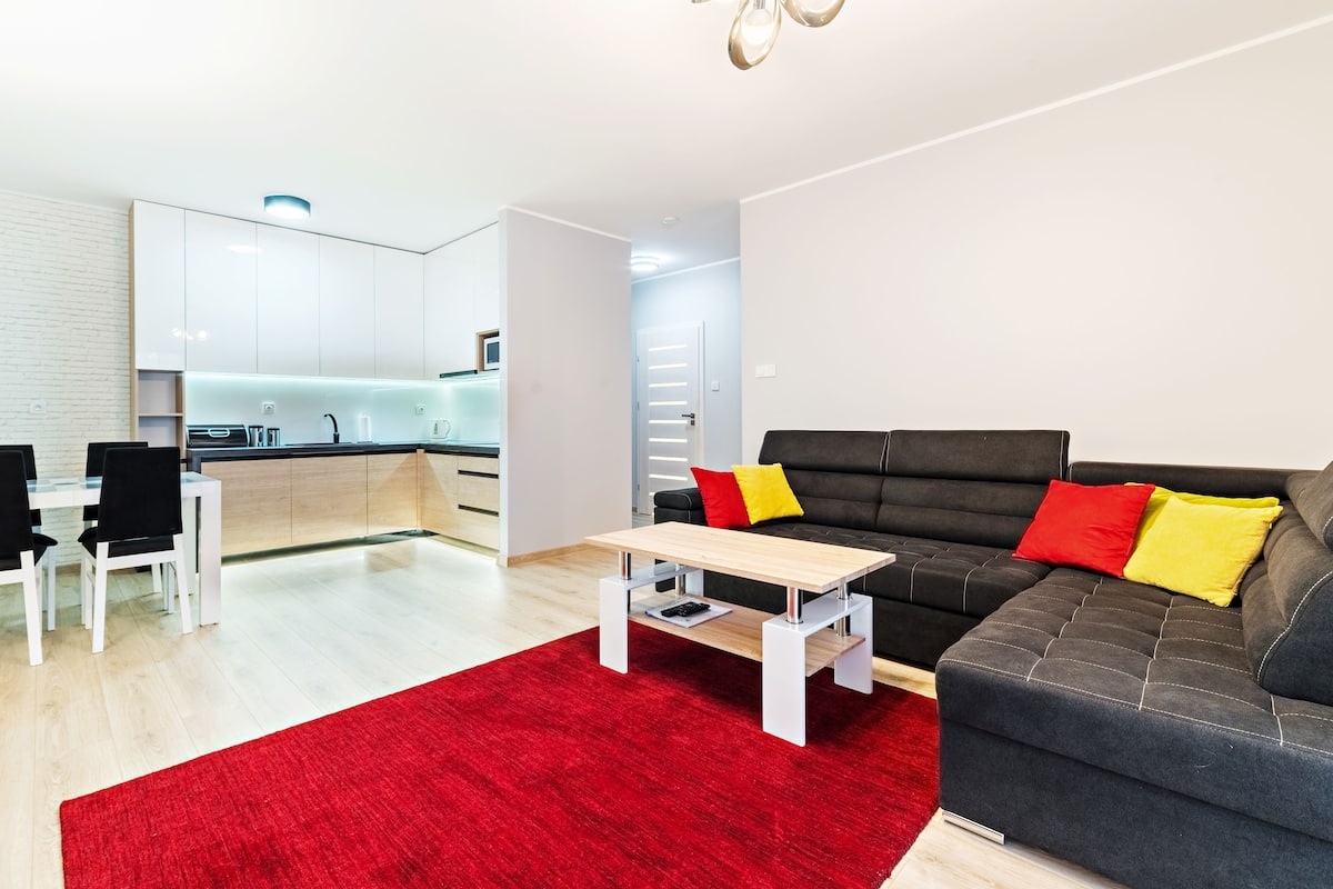 A modern living room with a black sofa, red and yellow cushions, a wooden coffee table, and a red rug.