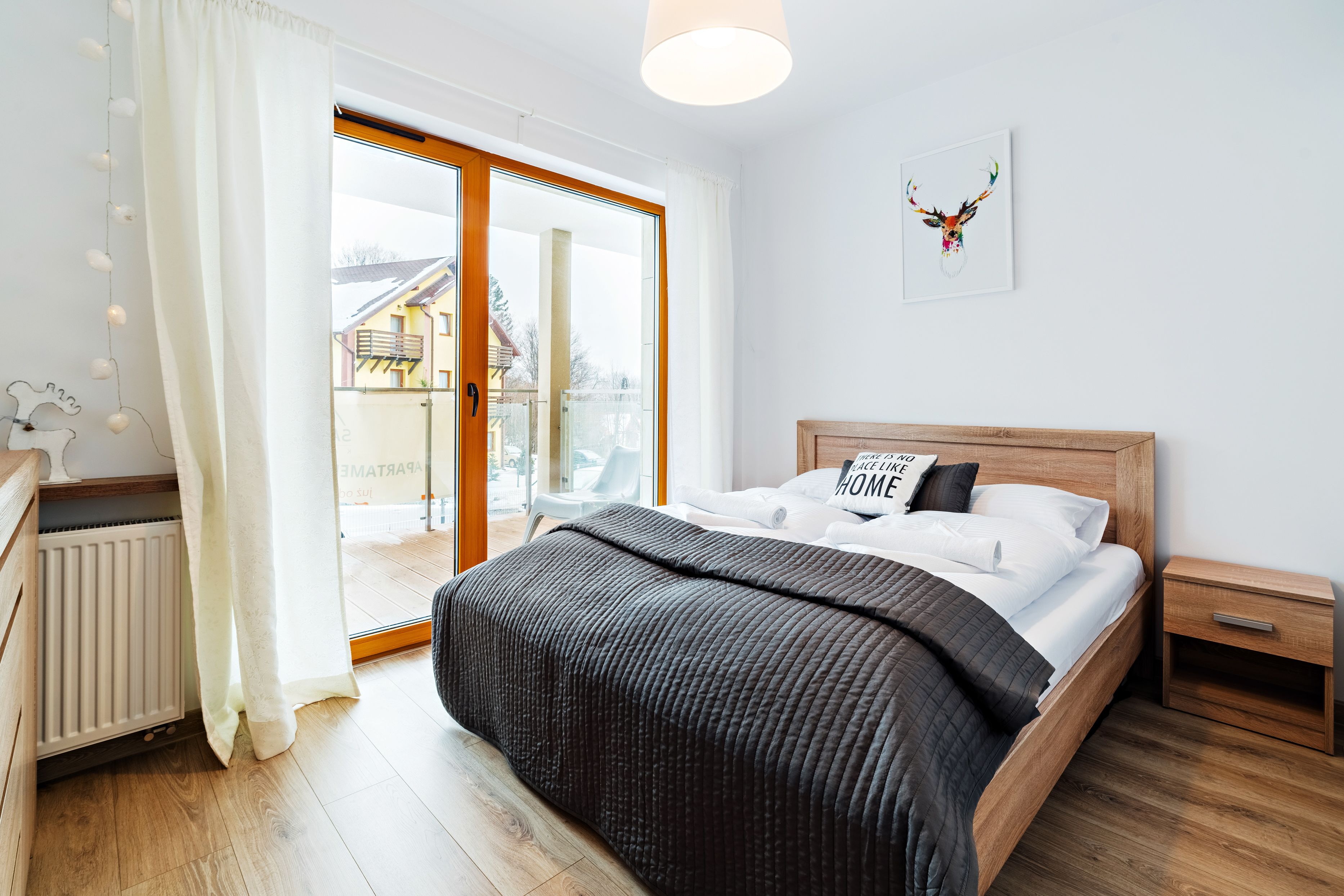A bedroom with a wooden bed, a nightstand, a radiator, and a view of a house through a glass door.