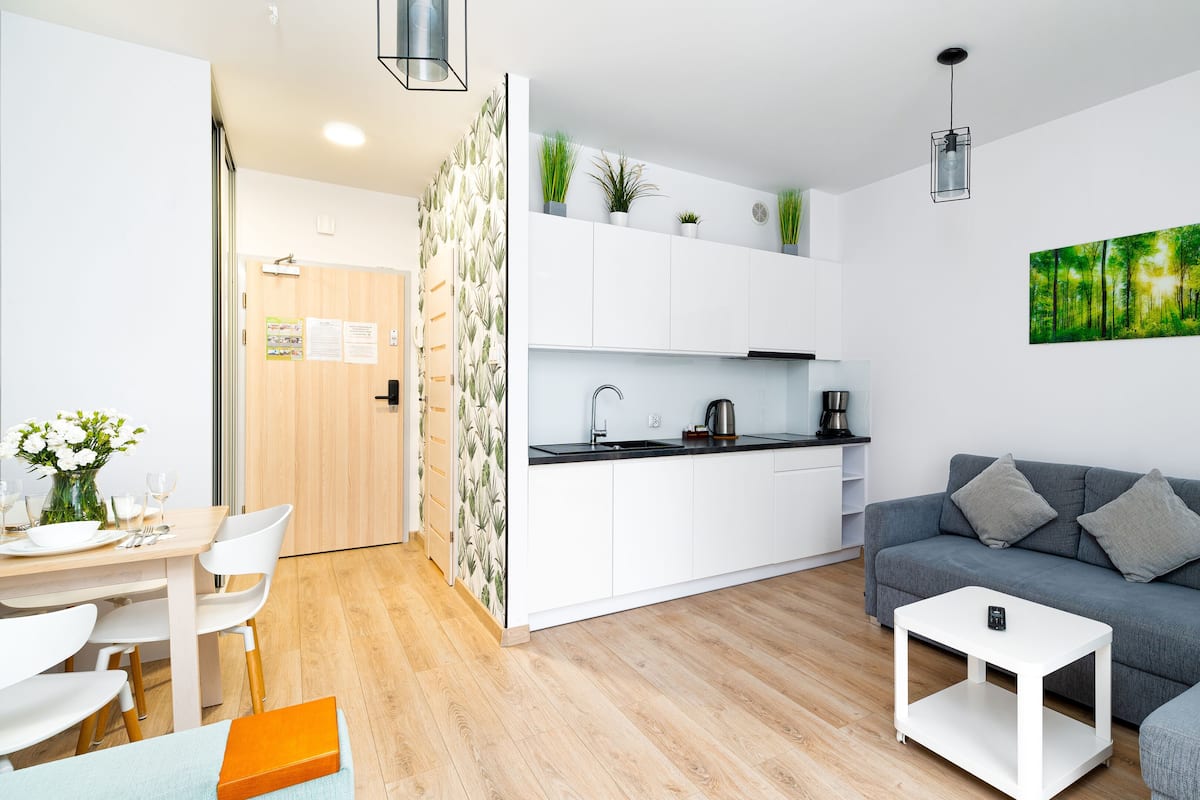 A modern living room with a grey sofa, a small white side table, and a dining area with a wooden table and chairs.