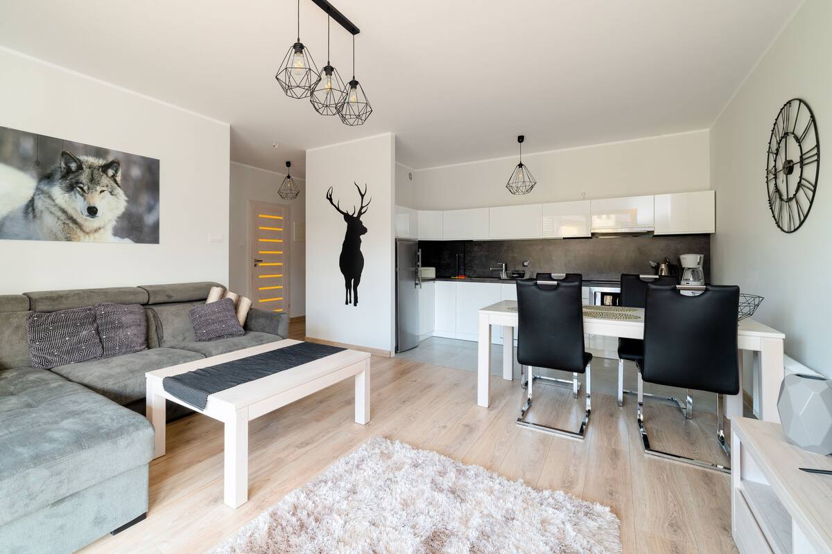 A modern living room with a grey sofa, patterned cushions, a coffee table, and a rug. A kitchen with white cabinets and a dining area with black chairs is visible in the background.