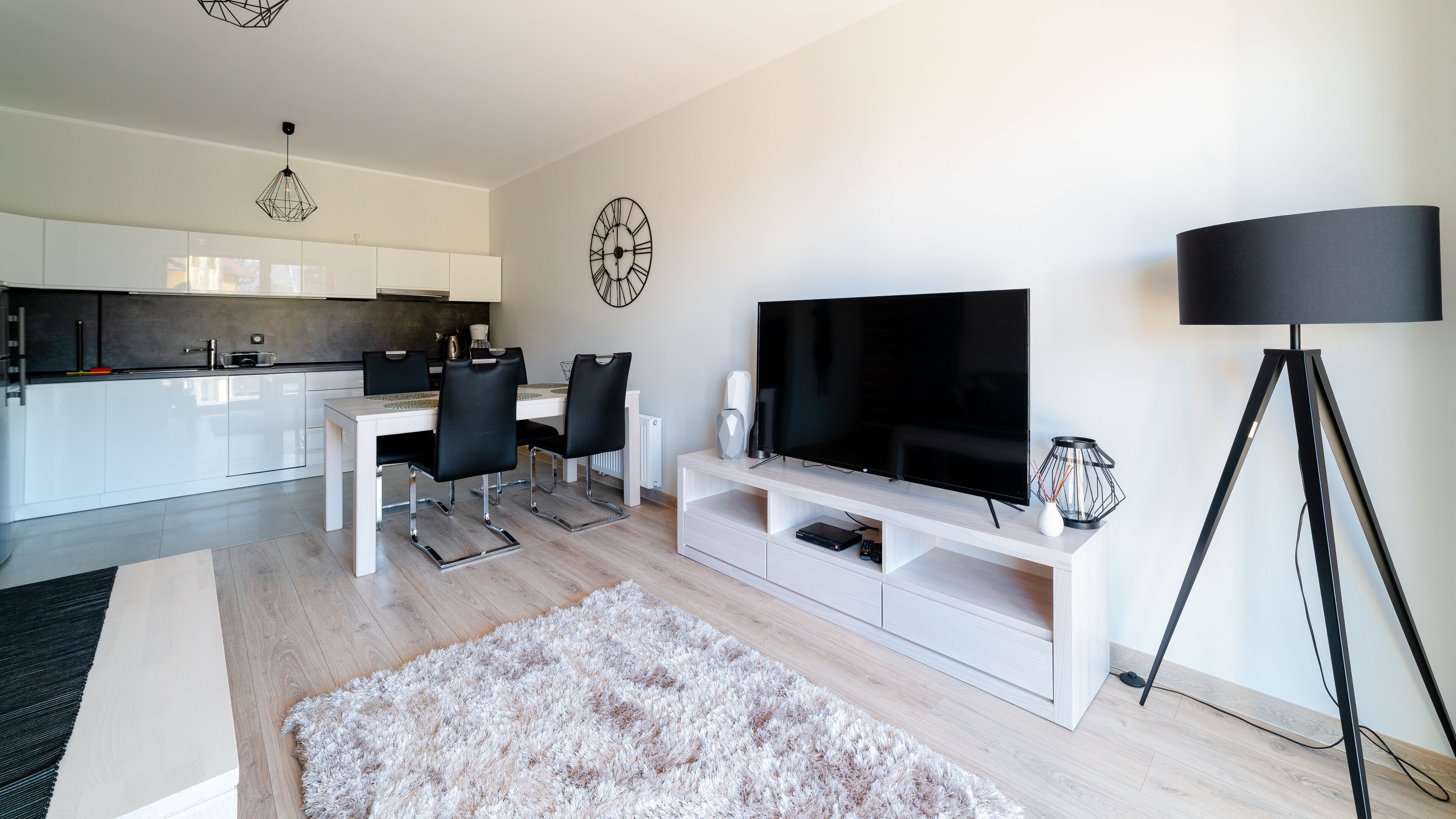 A modern living room with a flat-screen TV, a dining area with a white table and black chairs, and a kitchen in the background.