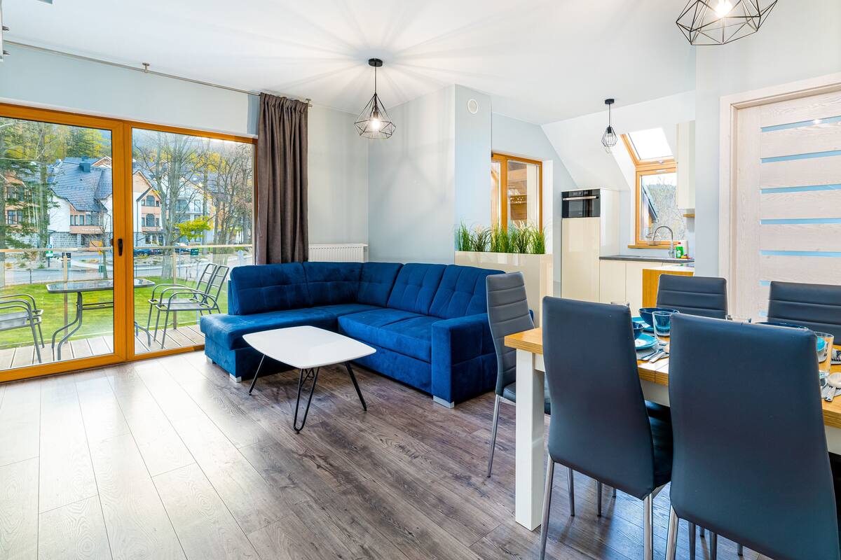 A modern living room with a blue sectional sofa, a dining table with chairs, and a view of the outside.
