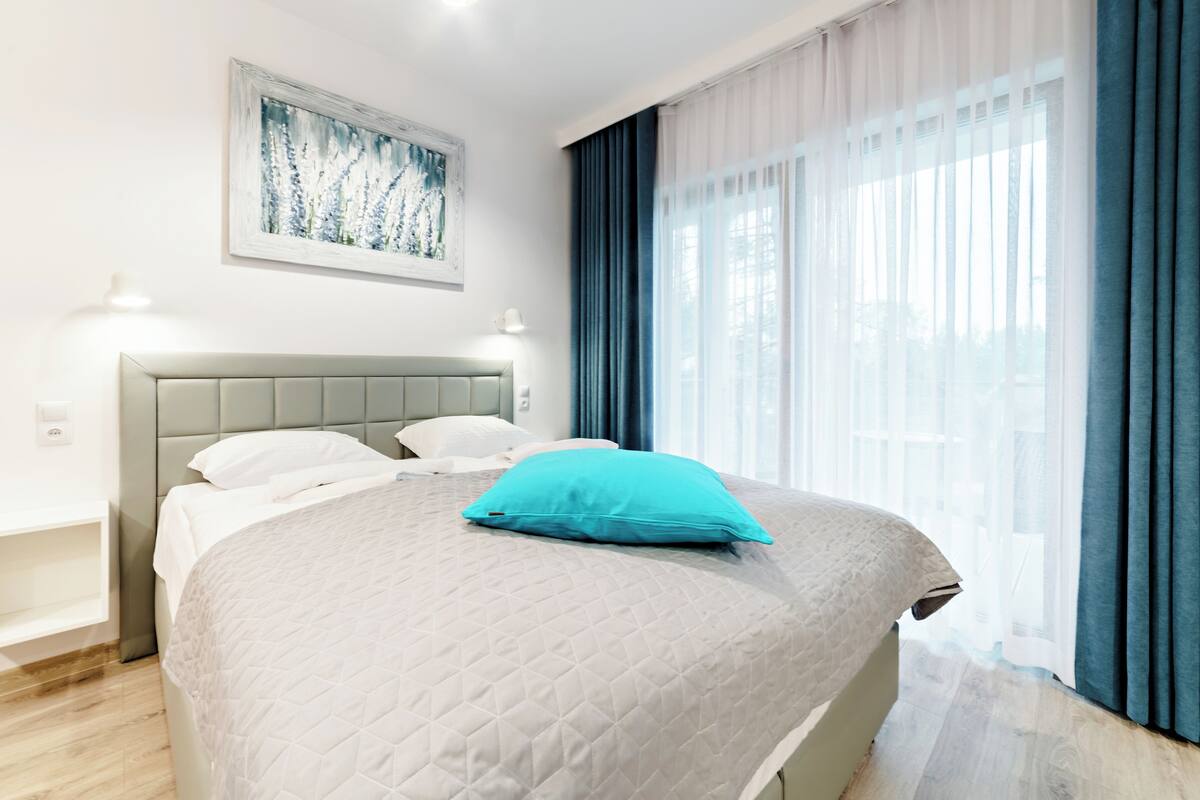 A modern hotel room with a large bed, a headboard, a painting on the wall, and a window with curtains.
