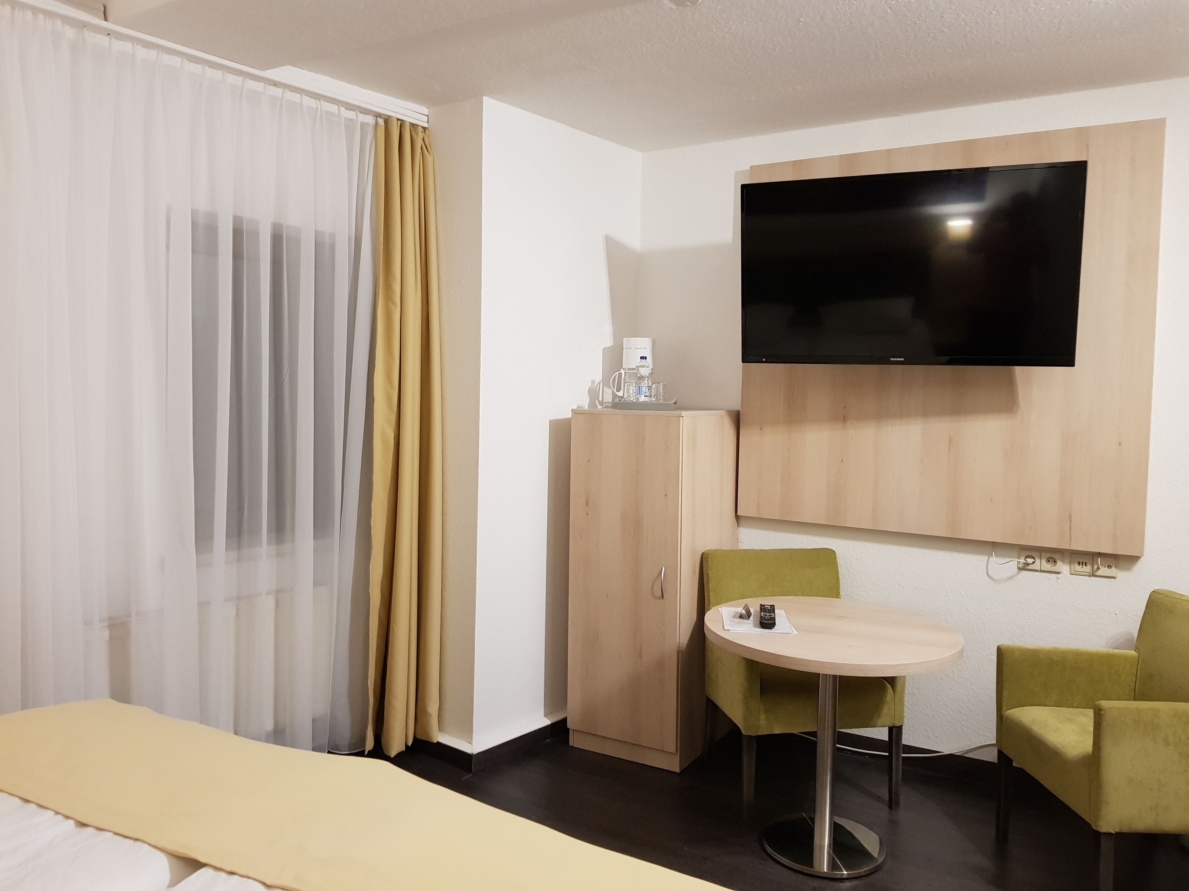 A hotel room with a bed, a desk with a chair, a television mounted on the wall, and a wardrobe.
