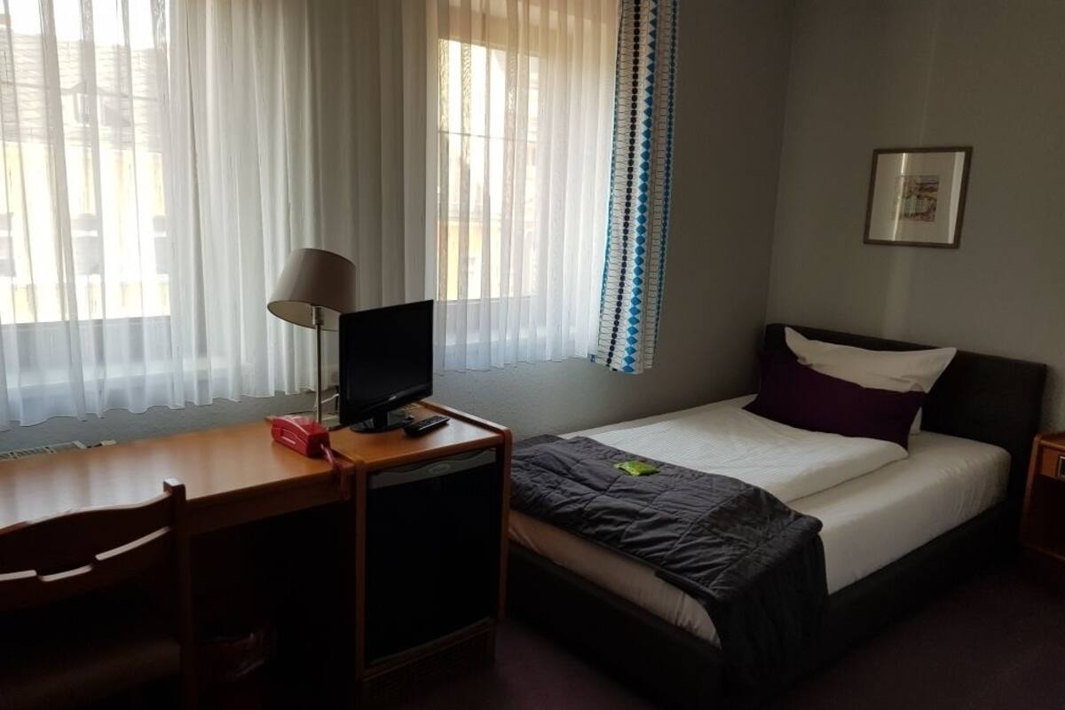 A bedroom with a bed, a desk with a computer, a chair, and a window with sheer curtains.