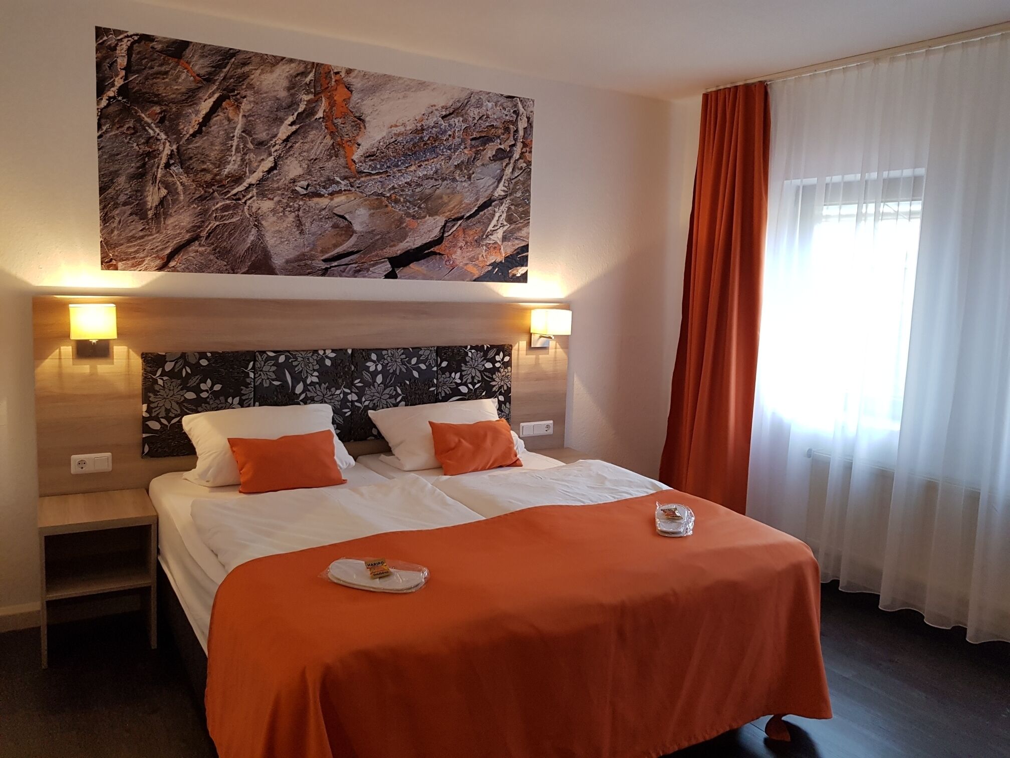 A hotel room with a bed, two nightstands, a marble artwork above the bed, and a window with curtains.