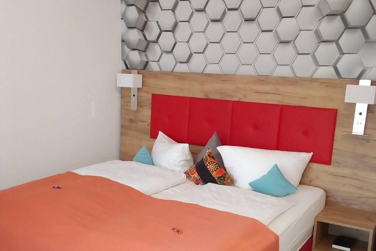 A modern bedroom with a wooden headboard, a geometric wall design, and a neatly made bed with orange bedding.