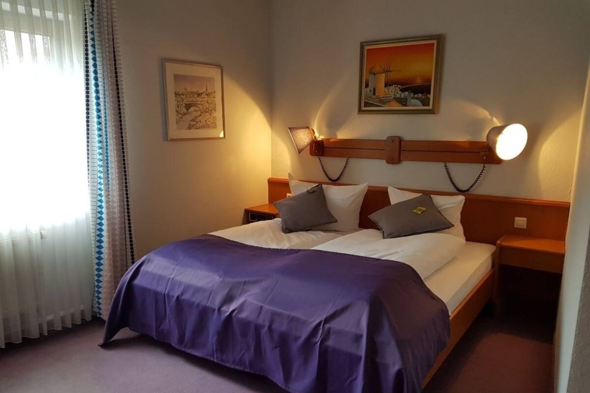 A hotel room with a bed, a nightstand, a lamp, a painting, and a window with curtains.