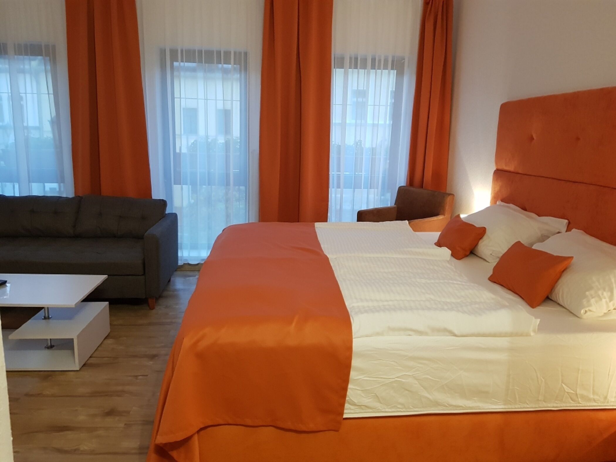 Ein Hotelzimmer mit einem großen Bett, orangefarbenen Vorhängen, einem grauen Sofa und einem weißen Couchtisch.