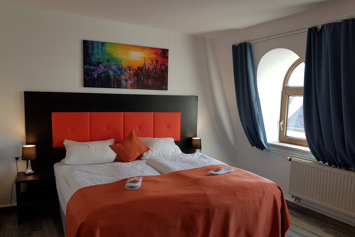 A hotel room with a large bed, a cityscape painting above the bed, a window with curtains, and a nightstand with a lamp.
