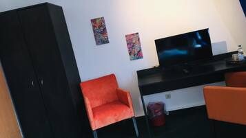 A modern room with a black desk, a flat-screen TV, two colorful paintings, and two orange chairs.